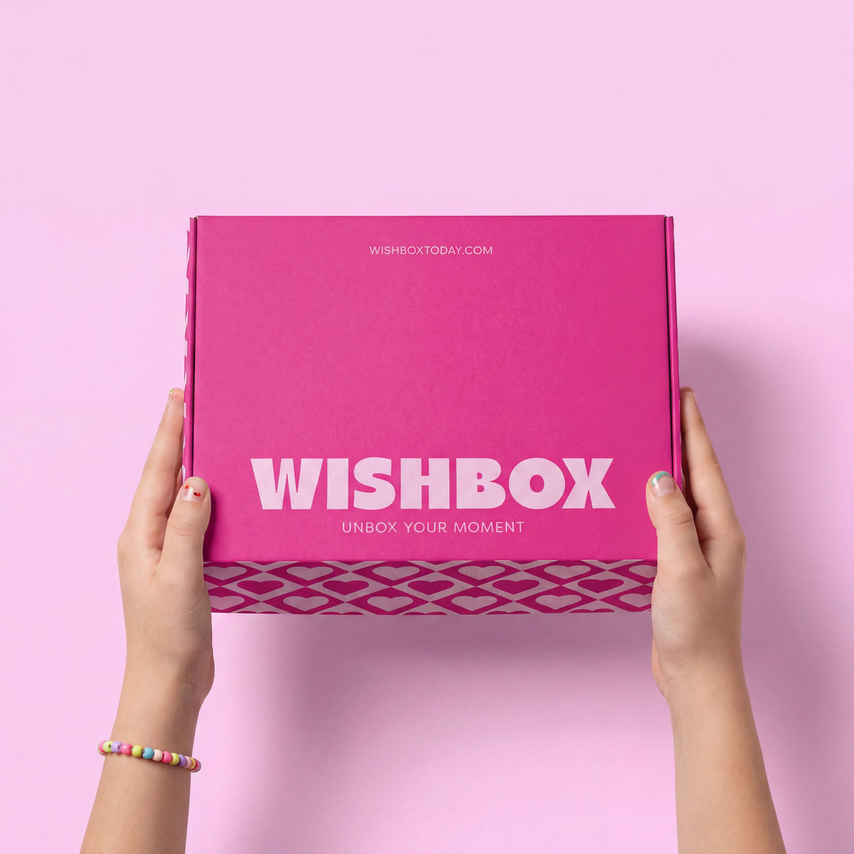 A person holds a bright pink Wishbox box against a solid pink background. The box features the white 'WISHBOX' logo and 'UNBOX YOUR MEMENT' text, with a patterned band at the bottom.