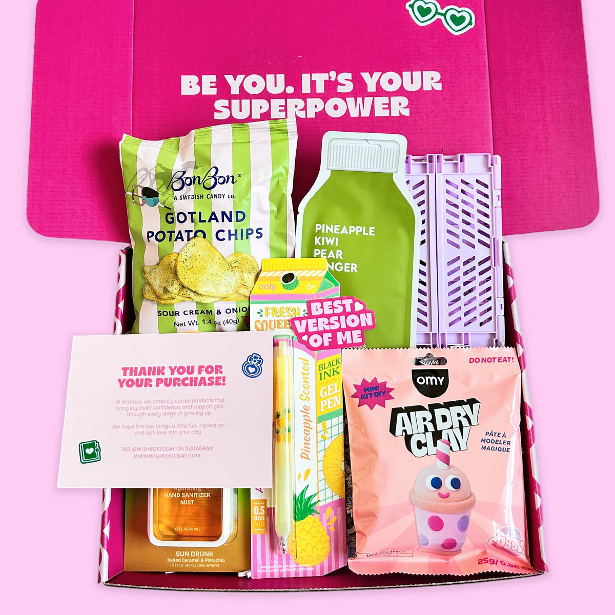 A curated snack and treat gift box arranged in a pink box with the lid open, featuring BonBon potato chips, a green pineapple kiwi finger candy, a pink Air Dry Clay bag, a yellow and pink 'Fresh Best of Version of Me' candy, a small bottle of honey, and a thank-you card. The box lid has the text 'BE YOU.